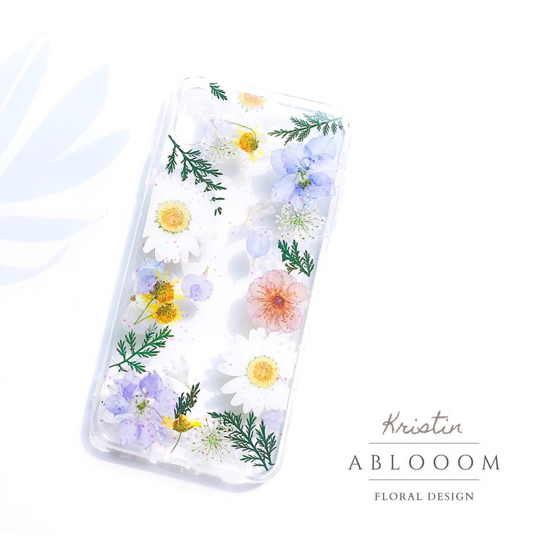 May include: A clear phone case with pressed flowers and greenery inside. The flowers are white, pink, blue, and yellow. The text "Kristin ABLOOOOM FLORAL DESIGN" is printed on the bottom of the case.
