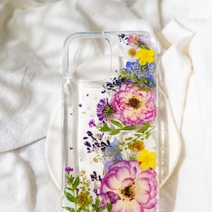 May include: A clear phone case with pressed flowers inside. The flowers are pink, purple, yellow, and blue. The case is designed for a smartphone with a camera bump.