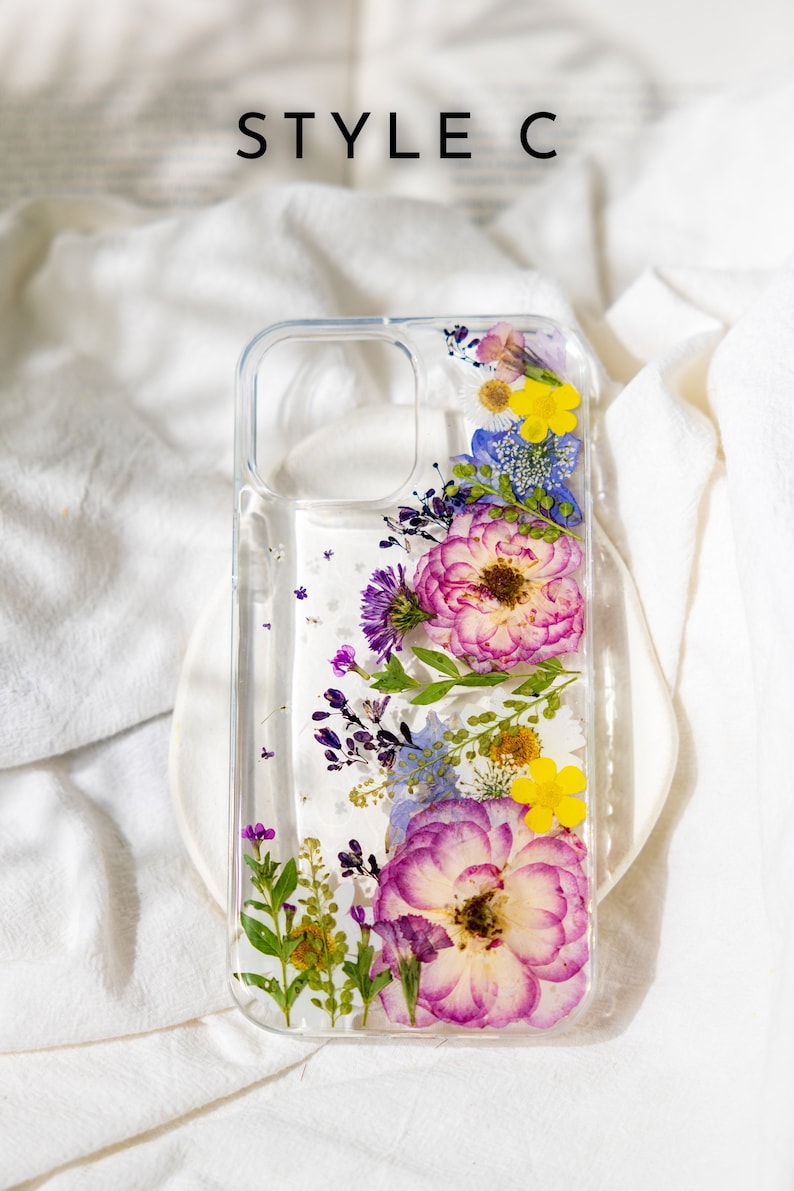 May include: A clear phone case with pressed flowers inside. The flowers are pink, purple, yellow, and blue. The case is designed for a smartphone with a camera bump.
