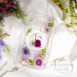 Könnte beinhalten: Eine transparente Handyhülle mit weißem Hintergrund und einem weiblichen Symbol. Die Hülle ist mit gepressten Blumen in den Farben Lila, Rosa und Weiß verziert. Der Text "Kristin ABLOOM FLORAL DESIGN" ist auf der Unterseite der Hülle aufgedruckt.