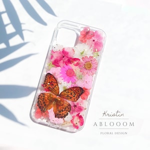 May include: A clear phone case with a pressed flower and butterfly design. The case features pink and white flowers, a brown butterfly, and gold glitter. The text "Kristin ABLOOOOM FLORAL DESIGN" is printed on the bottom of the case.