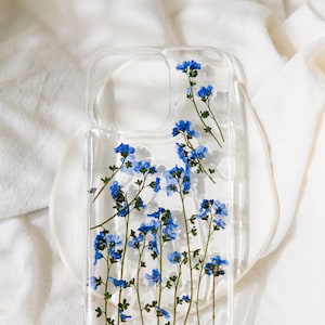 May include: A clear phone case with a design of pressed blue flowers. The case has a cutout for the camera lens.