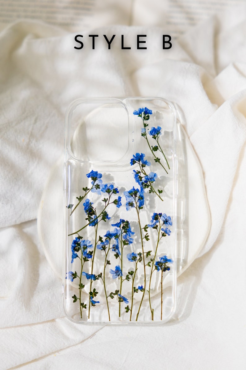 May include: A clear phone case with a design of pressed blue flowers. The case has a cutout for the camera lens.