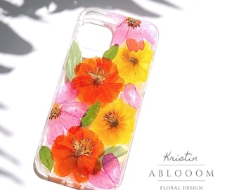 May include: A clear phone case with pressed flowers. The flowers are pink, orange, and yellow. The case has a white background with the text "Kristin ABLOOOOM FLORAL DESIGN".