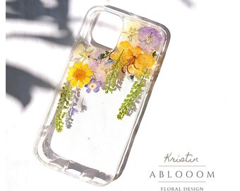 May include: A clear phone case with pressed flowers inside. The flowers are yellow, purple, and green. The case is designed by Kristin ABLOOOOM FLORAL DESIGN.