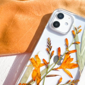 May include: A clear phone case with a pressed flower design. The flowers are orange and yellow and are arranged in a cluster. The case is designed for a phone with a dual camera lens.