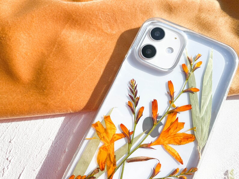 May include: A clear phone case with a pressed flower design. The flowers are orange and yellow and are arranged in a cluster. The case is designed for a phone with a dual camera lens.