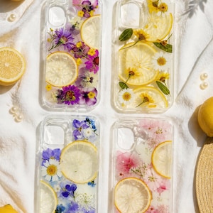 Può includere: Quattro cover per telefono trasparenti con fiori seci incorporati e fette di limone. La cover in alto a sinistra presenta fiori viola e gialli, quella in alto a destra fiori gialli, in basso a sinistra fiori blu e bianchi, e in basso a destra fiori rosa.