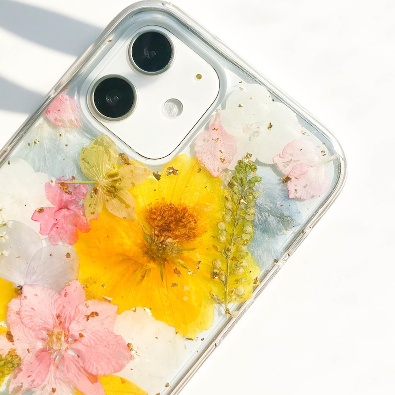 May include: A clear phone case with pressed flowers and gold leaf accents. The case features a yellow flower, pink flowers, blue flowers, and green leaves. The phone case is designed for a phone with a dual camera lens.
