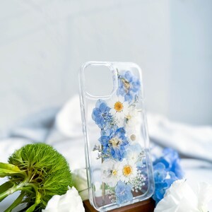 May include: A clear phone case with a pressed flower design. The case features blue and white flowers, including daisies and forget-me-nots.