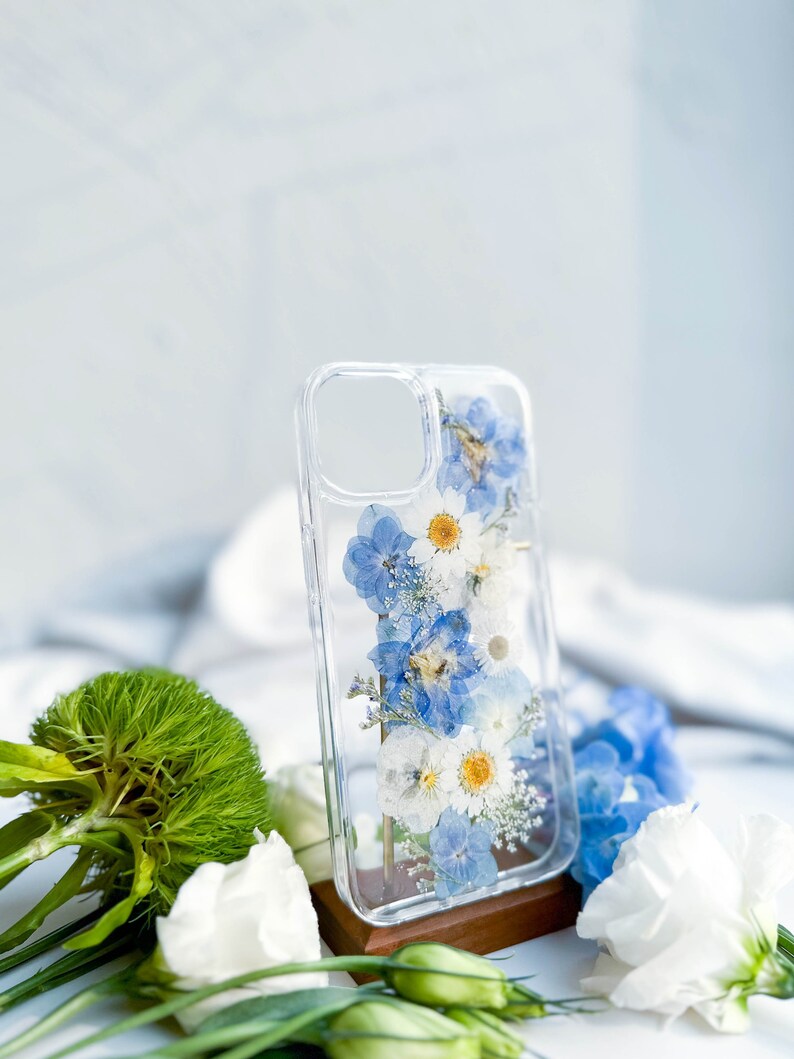 May include: A clear phone case with a pressed flower design. The case features blue and white flowers, including daisies and forget-me-nots.