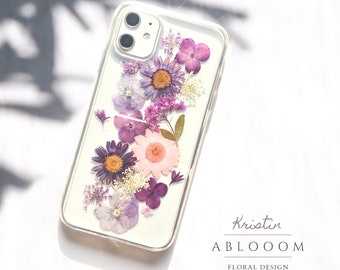 Könnte beinhalten: Eine transparente Handyhülle mit einem Blumendesign aus gepressten lila und rosa Blüten. Die Hülle hat einen Kameraausschnitt und ist für ein iPhone konzipiert. Der Text "Kristin ABLOOOOM FLORAL DESIGN" ist auf der Unterseite der Hülle aufgedruckt.