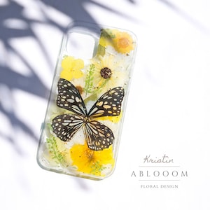 May include: A clear phone case with a pressed flower and butterfly design. The case features yellow and white flowers, green leaves, and a black and white butterfly. The text "Kristin ABLOOOOM FLORAL DESIGN" is printed on the bottom of the case.