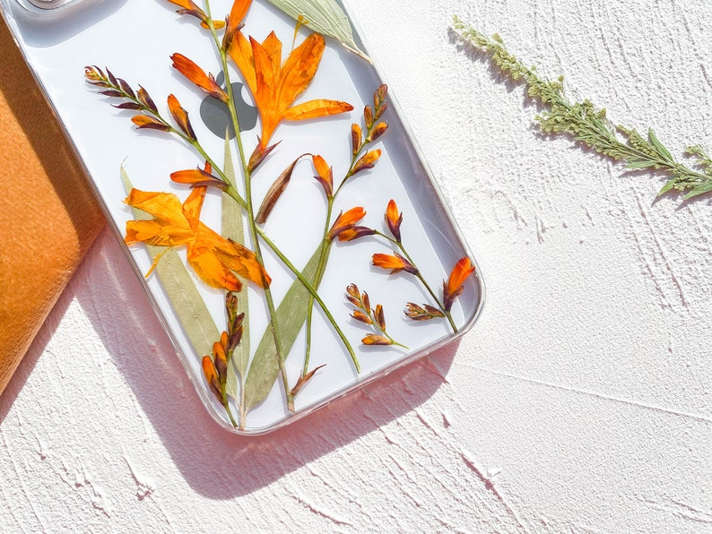 May include: A clear phone case with pressed orange and yellow flowers. The flowers are pressed between the clear plastic and the white phone case.