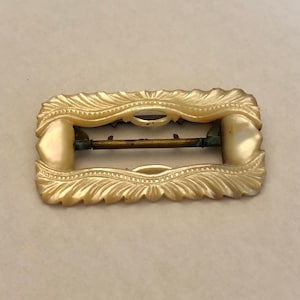 Peut inclure: Une boucle de ceinture vintage en nacre blanc cassé avec un design décoratif et orné. La boucle a une barre en laiton qui traverse le centre.