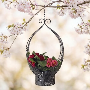 Könnte beinhalten: Ein schwarzer Metall-Hängekorb gefüllt mit roten und rosa Blumen und grünen Blättern. Der Korb hat ein dekoratives durchbrochenes Design und ist an einer Kette aufgehängt. Der Hintergrund zeigt unscharfe Kirschblütenzweige.
