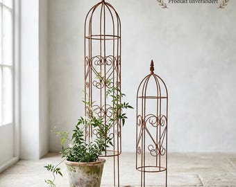 Op de afbeelding: Twee roestkleurige metalen tuinobelisken met verticale draadframes en decoratieve hartvormige krullen. Eén obelisk staat achter een verweerde terracotta pot met een groene klimplant, met een klein schepje ernaast.