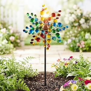 Könnte beinhalten: Ein farbenfroher Garten-Windspinner mit einer zentralen Bronzekugel und ausstrahlenden Armen aus bunten Kugeln. Der Windspinner steht in einem Gartenbeet mit grünen Pflanzen und Blumen, im Hintergrund ein Ziegelweg.
