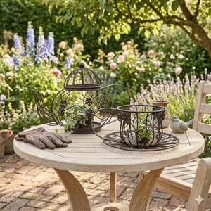 Könnte beinhalten: Ein dekoratives Metall-Teekannen- und Tassen-Pflanzgefäß-Set mit aufwendigen Blumendesigns, gefüllt mit Grün, steht auf einem Holztisch. Ein Paar braune Lederhandschuhe und eine kleine Vogelfigur befinden sich ebenfalls auf dem Tisch, mit Gartenhintergrund.