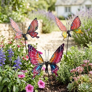 Puede incluir: Tres estacas de jardín de metal con forma de mariposa. Las mariposas tienen alas rojas, naranjas y azules con cuerpos y antenas negras. Están colocadas entre flores y vegetación en un jardín.