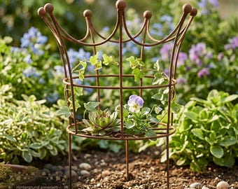 Op de afbeelding: Een roestkleurige metalen plantensteun in de vorm van een kroon met verticale staven en een ronde basis. De structuur bevat een kleine groene vetplant en een wit met paars viooltje, met groene klimopranken door het frame.