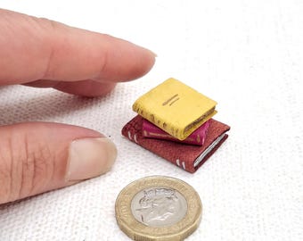 May include: A stack of three miniature books in shades of yellow, pink, and burgundy. The books are stacked on top of each other. A gold coin is next to the books. The books are small, likely for a dollhouse or miniature collection.