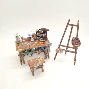 Op de afbeelding: Een miniatuur kunstenaarsatelier-set bestaande uit een houten werktafel met schildersbenodigdheden, penselen en kleine doeken, een bijpassend houten krukje met een beschilderd bloemendoek en een hoge houten ezel met verfpalet.