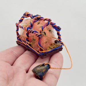 May include: A miniature orange fishing net cage with blue and orange rope detailing, set on a wooden base. A small, dark blue and orange weight is attached by a string. The cage is filled with moss and small objects.