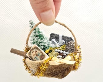 May include: Miniature Christmas basket filled with a small tree, a wrapped gift, pine cone, and a vintage card. The basket is trimmed with gold tinsel and a cream-colored bow. The handle is made of twisted twine.