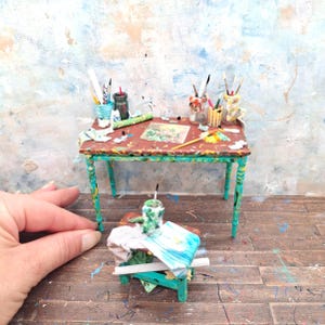Op de afbeelding: Miniatuur kunstenaarstafel met een roodbruin oppervlak en turquoise poten. De tafel is bedekt met verf, penselen en kunstbenodigdheden. Een kleinere bijpassende tafel staat ervoor. De scène speelt zich af op een houten vloer.