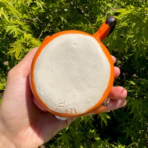 May include: A handmade ceramic mug with a fox design. The mug is orange with black accents and a white interior. The handle is shaped like a fox's tail. The bottom of the mug has the word "yin" written on it.