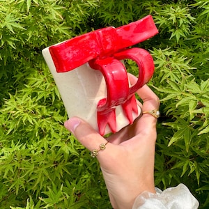 May include: A ceramic mug with a red bow handle and bow detail. The mug is off-white with speckles. The bow handle is a bright red color. The mug is held in front of a green leafy background.