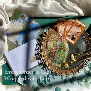 May include: An artistic flat lay featuring a decorative gold-rimmed mirror with tarot cards and stained glass stickers. An open envelope with a painting of a woman with an umbrella is visible. The text "Every Order is Gift Wrapped with Love" is at the bottom.
