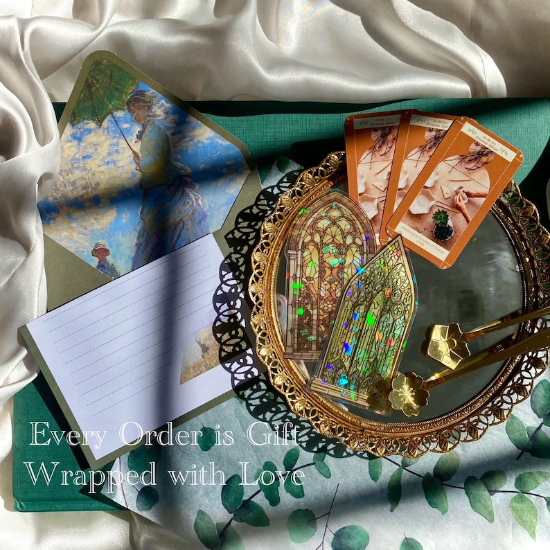 May include: An artistic flat lay featuring a decorative gold-rimmed mirror with tarot cards and stained glass stickers. An open envelope with a painting of a woman with an umbrella is visible. The text "Every Order is Gift Wrapped with Love" is at the bottom.