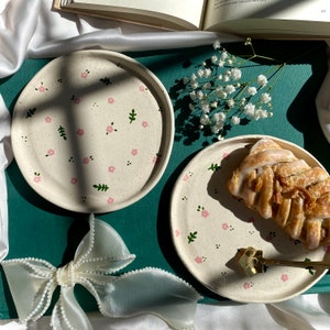 May include: Two white ceramic plates with a floral pattern in pink and green. The plates are on a green surface. One plate has a pastry on it.