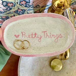 May include: Oval ceramic trinket dish with a pink border and speckled interior. The dish has the words "Pretty Things" in pink script, with a pink heart. Two gold rings are inside the dish.