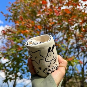 May include: A cream-colored ceramic tumbler with black line art of a cartoon character on a skateboard, a star, and other shapes. The tumbler has a cylindrical shape with a slightly flared top. The background is a blurred view of a tree with colorful autumn leaves.
