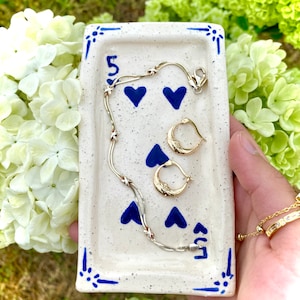 May include: A white ceramic trinket dish with a blue and white design featuring hearts and the number "5". A gold chain necklace and a pair of gold hoop earrings are displayed on the dish.