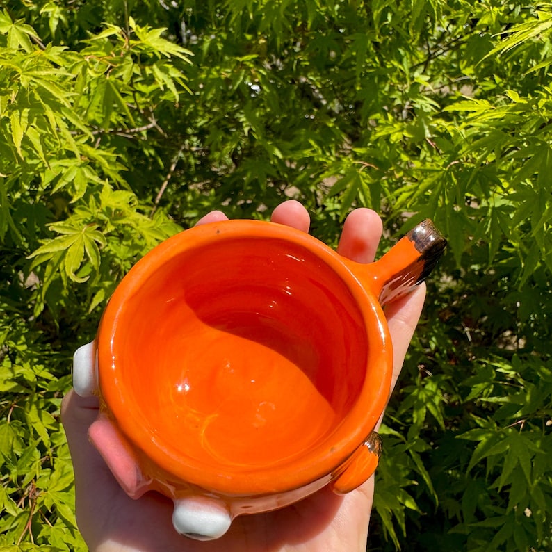 May include: Bright orange ceramic bowl with a handle, white and pink accents, and a brown-tipped handle. The bowl is held in front of a background of green leaves. The bowl is likely handmade.