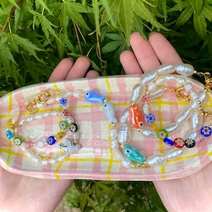 May include: Two pearl necklaces with colorful glass beads and charms, including fish and a dove, displayed on a pink and yellow checkered plate.