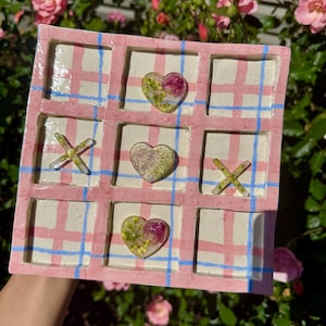 May include: A handmade tic-tac-toe game board. The board is square, with a pink and white plaid pattern and blue grid lines. The game pieces are clear resin hearts and X's, each containing dried flowers. The board is held by a person.