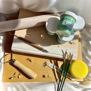 May include: A collection of pottery tools and supplies, including jars of green glaze, paintbrushes, a rolling pin, a sponge, and gold leaf flakes. The tools are arranged on a stack of paper and a book.