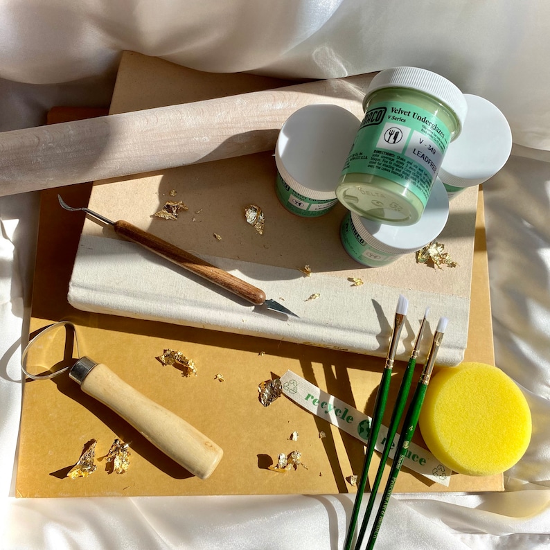 May include: A collection of pottery tools and supplies, including jars of green glaze, paintbrushes, a rolling pin, a sponge, and gold leaf flakes. The tools are arranged on a stack of paper and a book.