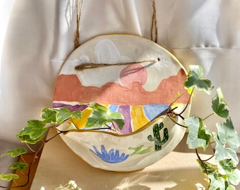 May include: A white ceramic hanging planter with a colorful painted landscape scene. The planter features a sunset, mountains, and a cactus. The planter is hanging from a twine rope.