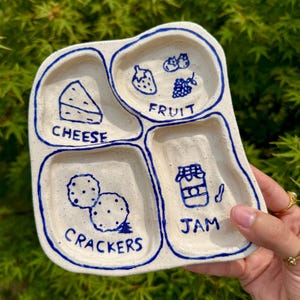 May include: A handmade, off-white ceramic serving dish with four compartments. Each section is outlined in blue and features a hand-drawn food illustration and label: cheese, fruit, crackers, and jam. The dish is held against a backdrop of green foliage.