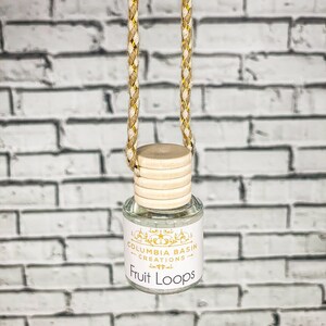 Puede incluir: Una pequeña botella de vidrio transparente con tapa de madera y colgador de cuerda trenzada. La etiqueta dorada dice "Columbia Basin Creations" y "Fruit Loops". El fondo es una pared de ladrillos blancos.