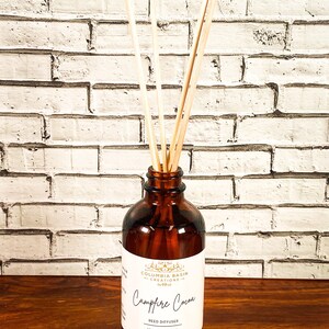 Puede incluir: Un difusor de caña de vidrio marrón con cañas de madera. La etiqueta dice "Campfire Cocoa" y "100% Natural". La botella contiene 120 ml de líquido. El fondo es una pared de ladrillos blancos.