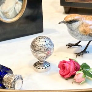 May include: An antique still life arrangement featuring a silver salt shaker with ornate detailing, a cobalt blue bottle, a decorative bird figurine, a framed portrait, and pink roses. The objects are arranged on a white surface.