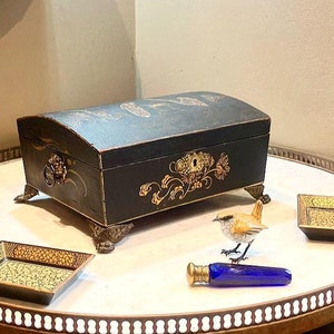 May include: A black wooden box with gold painted details and a lion head handle. The box is decorated with gold painted flowers and leaves. The box is sitting on a table with a small bird figurine and a tray.