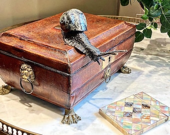 May include: A vintage leather-bound box with brass lion head accents and feet. The box has a brass lock and keyhole. A decorative snail sculpture is perched on top of the box.